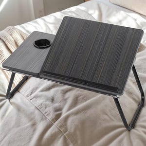 May include: A portable laptop desk with a dark wood grain finish, featuring a tilting surface with a black ledge and a flat side section with a circular cup holder. The desk is supported by thin, black foldable metal legs on a flat surface.