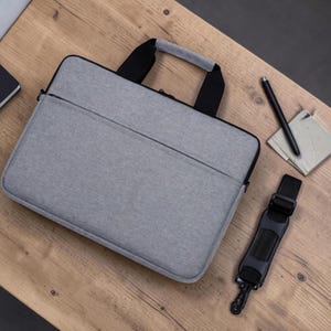 May include: A rectangular grey laptop bag with black trim and black handles. The bag features a front zipped pocket and a detachable black shoulder strap with a padded section. The design is simple and functional with a textured fabric finish.