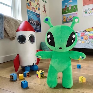 Può includere: Un peluche alieno verde brillante con grandi occhi neri e antenne, in piedi accanto a un razzo giocattolo bianco e rosso. Diversi piccoli cubi colorati sono sparsi sul pavimento attorno al peluche alieno e al razzo giocattolo.