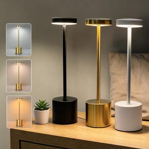 May include: Three cordless table lamps with cylindrical bases and thin stems, featuring flat circular light heads. The lamps are finished in matte black, brushed gold, and white. Each lamp emits a soft downward glow, showcasing three distinct color temperatures.