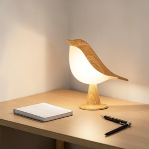May include: A bird-shaped table lamp with a light-colored wood grain finish on the head, back, and base, featuring a glowing white translucent body that emits a soft light. The lamp stands on a circular base and is positioned on a flat surface.