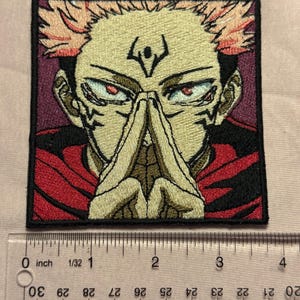 Pu&ograve; includere: Una toppa ricamata quadrata raffigurante un personaggio anime stilizzato con capelli a punta color pesca, occhi rossi e segni scuri sul viso. Il personaggio indossa un indumento rosso e tiene le mani giunte. Un righello indica 8,9 cm.