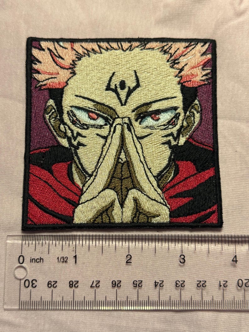 Pu&ograve; includere: Una toppa ricamata quadrata raffigurante un personaggio anime stilizzato con capelli a punta color pesca, occhi rossi e segni scuri sul viso. Il personaggio indossa un indumento rosso e tiene le mani giunte. Un righello indica 8,9 cm.