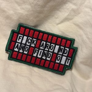 May include: A rectangular embroidered patch with a dark green border and a red grid pattern. The patch features white square tiles with black letters spelling out the phrase FUCK AROUND AND FIND OUT, arranged in two rows across the red grid.