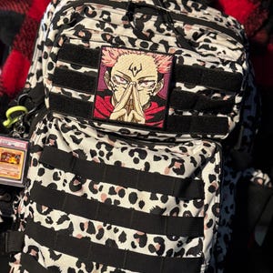 Pu&ograve; includere: Uno zaino tattico con motivo leopardato bianco, nero e rosa chiaro, dotato di cinghie MOLLE nere. Una toppa quadrata ricamata raffigurante un personaggio anime con capelli rosa e occhi rossi &egrave; fissata sulla tasca anteriore.