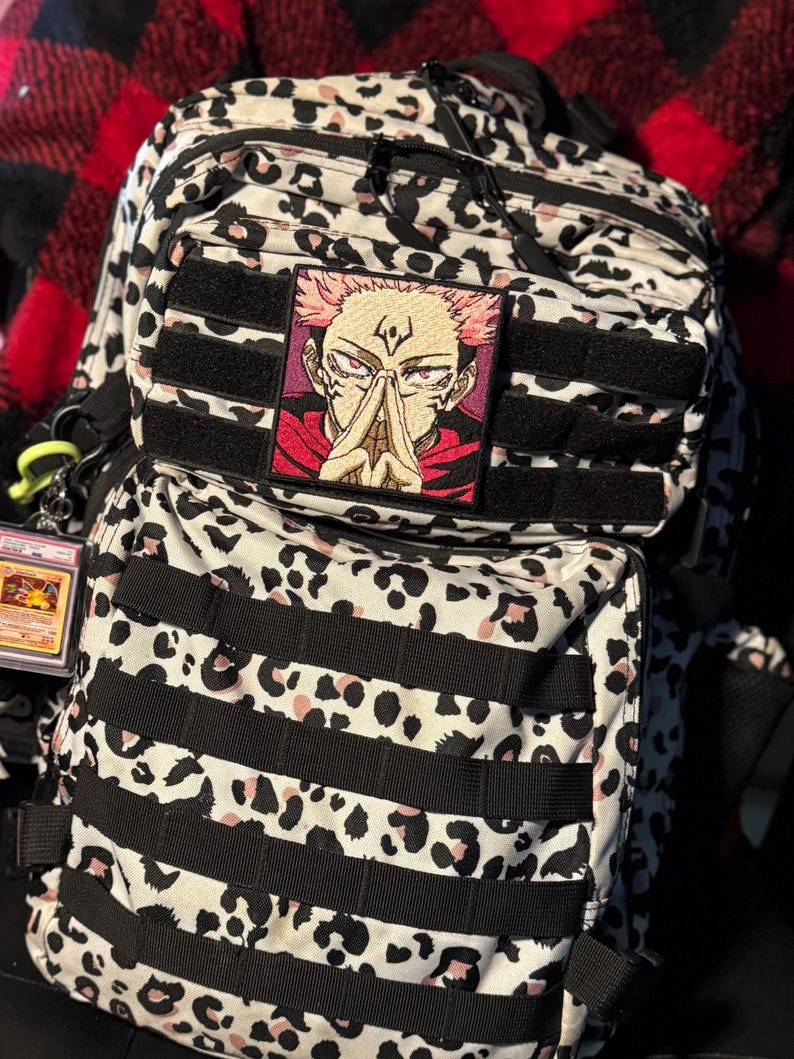 Pu&ograve; includere: Uno zaino tattico con motivo leopardato bianco, nero e rosa chiaro, dotato di cinghie MOLLE nere. Una toppa quadrata ricamata raffigurante un personaggio anime con capelli rosa e occhi rossi &egrave; fissata sulla tasca anteriore.