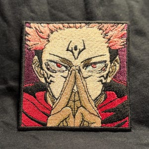 May include: A square embroidered patch featuring a stylized anime character with spiky pink hair, red eyes, and a black forehead marking. The character is shown with hands pressed together in a prayer-like gesture, wearing a red and black garment.