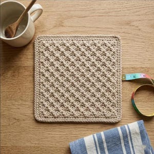Könnte beinhalten: Ein quadratischer, beigefarbener Häkeluntersetzer mit strukturiertem Rautenmuster. Ein Maßband liegt an der Seite und zeigt eine Breite von etwa 12,7 cm an. Eine weiße Keramiktasse mit Holzlöffel und ein blau-weiß gestreiftes Tuch liegen daneben.
