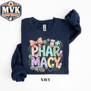 May include: A navy blue crewneck sweatshirt featuring a colorful graphic print that spells out PHARMACY in various pastel patterns. The design includes a plaid bow, a mortar and pestle, a pill bottle, a stethoscope, and small floral accents.