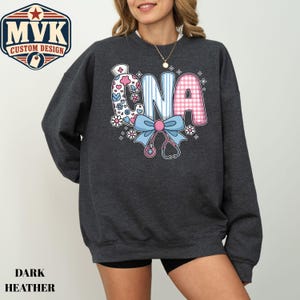 May include: A dark heather gray crewneck sweatshirt featuring a graphic print of the letters CNA. The C is decorated with flowers, the N has blue stripes, and the A has a pink gingham pattern. A light blue bow and a stethoscope are below the letters.