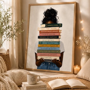 Op de afbeelding: Een ingelijste muurkunstprint met een persoon met donker haar in een knot die een stapel kleurrijke boeken voor het gezicht houdt. De boekruggen bevatten teksten zoals Growing & Glowing, Focus Plan Succeed, Discipline Creates Freedom en I Can & I Will.