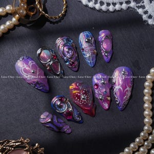 May include: A set of ten press-on nails featuring a purple and blue gradient base with intricate silver metallic swirls, embedded rhinestones, and star-shaped charms. The designs include plaid patterns, heart motifs, and iridescent finishes.