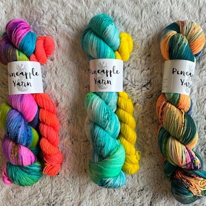 May include: Three skeins of colorful Pineapple Yarn. The yarn is dyed in vibrant hues of pink, blue, green, orange, and yellow. Each skein has a white label with the words "Pineapple Yarn" printed on it. The yarn is arranged on a textured, light-colored surface.