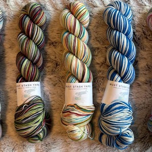 May include: Five skeins of Must Stash Yarn in various colorways. The yarn is a mix of colors, including blue, green, red, and yellow. Each skein has a label with the brand name and the word "Eras". The yarn is arranged on a textured surface.