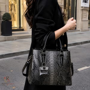 Puede incluir: Un bolso estructurado con un patrón de piel de serpiente en negro y gris con acabado brillante. El bolso incluye dos asas superiores, una correa de hombro desmontable y un pequeño dije a juego que cuelga de los herrajes frontales.