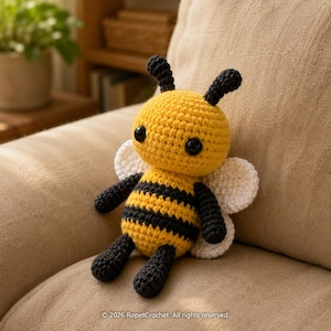 May include: A handmade crochet bee doll with a yellow body, black stripes, and black limbs. The bee has white wings and black antennae. The doll is sitting on a beige surface. The text "© 2026 RopetCrochet. All rights reserved." is at the bottom.