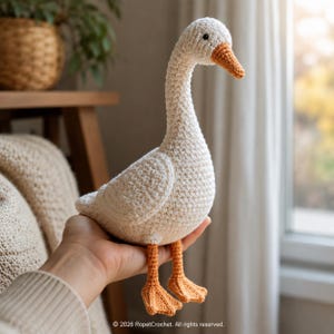 May include: A cream-coloured, crocheted goose with an orange beak and feet. The goose is held in a hand. The soft sculpture is likely a toy or decorative item. The goose is made by RopetCrochet.