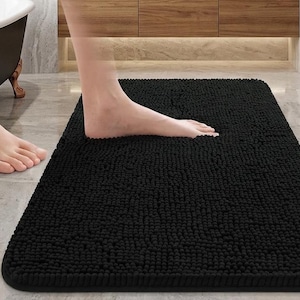 Puede incluir: Una alfombrilla de baño rectangular de color negro con una superficie de chenilla densa y texturizada que presenta un aspecto irregular similar a pequeños guijarros. La alfombra tiene un borde limpio y un color negro uniforme.
