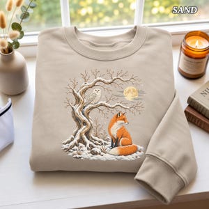 May include: A sand-colored crewneck sweatshirt featuring a graphic print of a snowy winter scene with a white owl perched on a bare tree branch above a red fox sitting in the snow under a full moon. The text SAND is printed in the top right corner.