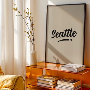 Könnte beinhalten: Ein gerahmter, rechteckiger Wandkunstdruck mit dem Wort Seattle in schwarzer Schreibschrift auf hellbeigem Hintergrund. Der Druck befindet sich in einem dünnen schwarzen Rahmen und steht auf einem orangefarbenen, durchscheinenden Tisch mit gestapelten Büchern.