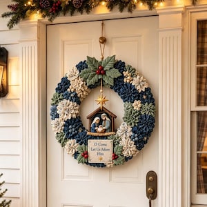 May include: A circular Christmas wreath featuring blue and white pinecones, green fabric holly leaves, and red berries. At the centre hangs a wooden nativity scene with a gold star above and a plaque below that reads O Come Let Us Adore Him.