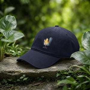 Op de afbeelding: Een marineblauwe baseballpet met een afbeelding van twee veren in geel- en blauwtinten, met het woord plume in witte letters onder de veren gedrukt. De pet heeft een gebogen klep en een gestructureerde kroon met gestikte oogjes.