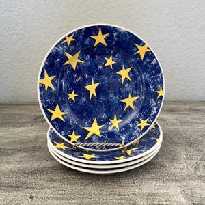 May include: A stack of four round ceramic plates featuring a deep blue background with a mottled white texture and scattered yellow five-pointed stars. The plates are held upright by a thin, twisted gold-toned metal wire display stand.