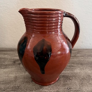 May include: A terracotta-colored ceramic pitcher featuring a rounded body, a curved handle, and a spout. The exterior is decorated with horizontal ribbed bands at the neck, wavy incised lines around the center, and dark brown drip-like accents.