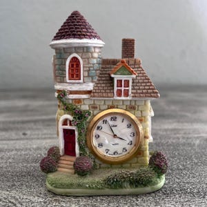 May include: A miniature house-shaped table clock featuring a stone-textured facade, a tall turret with a dark roof, and a red door. A round gold-rimmed clock face with black numerals and the text "Quartz" is embedded in the front wall.