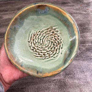 May include: A round ceramic grater plate featuring a light sage green glaze with brown and blue accents along the rim. The center of the plate has a raised, textured spiral pattern designed for grating food items like garlic or ginger.