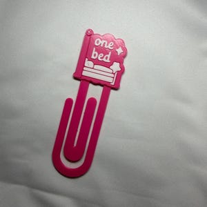 May include: A bright pink plastic paperclip bookmark featuring a decorative topper shaped like a bed with the white text one bed and two small star icons. The topper is attached to a large, elongated U-shaped paperclip design for page marking.