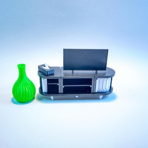 Pode incluir: Um móvel de televisão em miniatura preto com uma televisão de ecrã plano montada em cima e uma pequena caixa preta no lado esquerdo, acompanhado por um vaso bulboso verde brilhante e canelado à esquerda do móvel.