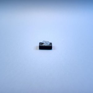 Pode incluir: Um pequeno bloco preto retangular com um acabamento liso e mate. Um único pedaço de película plástica transparente, fina e ligeiramente amarrotada, está fixado na superfície superior do bloco preto, perto do centro do objeto.