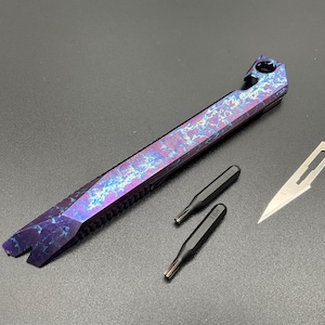 May include: A multi-tool pry bar featuring a vibrant purple and blue lightning-patterned finish, accompanied by two black screwdriver bits and a single silver-coloured scalpel blade. The pry bar includes a circular hole and a notch at one end.