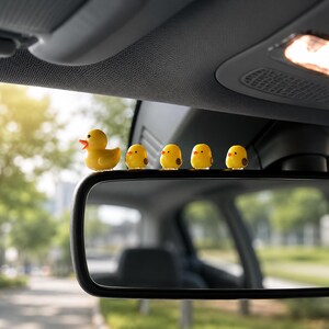 May include: A set of five small yellow figurines attached to the top edge of a car rearview mirror. The collection includes one larger yellow duck with an orange beak and four smaller yellow chicks, all positioned in a row facing forward.