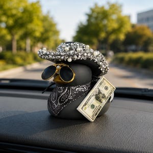 May include: A matte black rubber duck figurine wearing a wide-brimmed hat covered in silver rhinestones and white pearls. The duck features gold-rimmed round sunglasses, a black and white paisley bandana, and a stack of miniature ten-dollar bills.