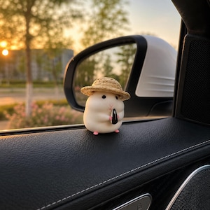 May include: A small, white, rounded figurine of a hamster wearing a tan straw hat. The hamster has black eyes, a pink nose, and tiny pink feet, and it is holding a single black sunflower seed between its front paws in a seated position.