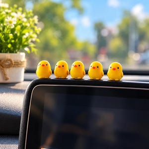 May include: Five small, bright yellow chick figurines with orange beaks and black eyes are lined up in a row along the top edge of a black vehicle dashboard display screen. Each miniature bird has a smooth, rounded body and a cheerful expression.