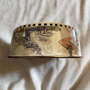 May include: A cream-colored metal cake dome with a decorative landscape scene featuring deer, trees, and a winding path. The dome has a gold rim and a checkered pattern around the top edge.