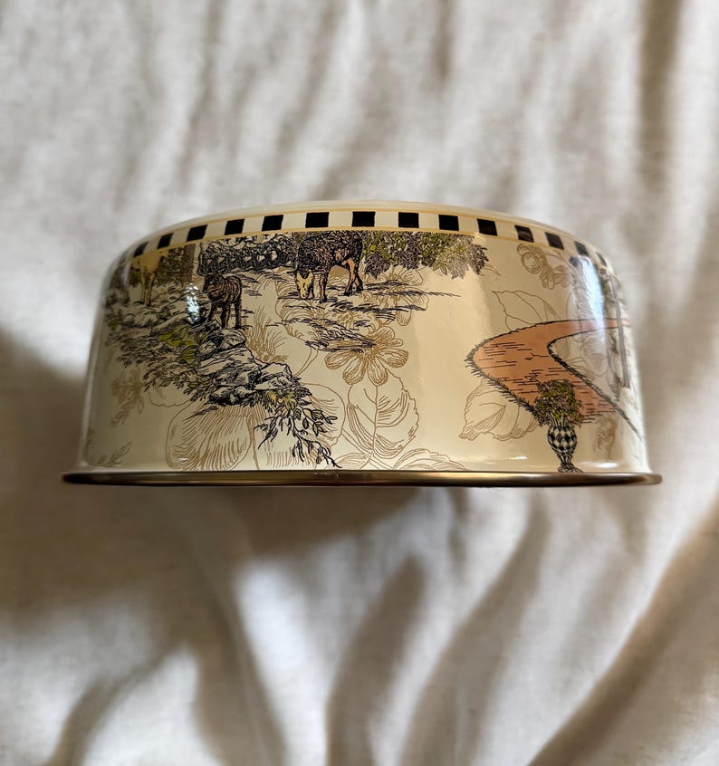 May include: A cream-colored metal cake dome with a decorative landscape scene featuring deer, trees, and a winding path. The dome has a gold rim and a checkered pattern around the top edge.