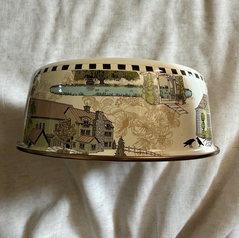 May include: A cream-colored, dome-shaped cake cover with a detailed landscape design. The illustration features houses, trees, and a pond, with a black and brown border. Two black birds are perched on the edge of the cover.
