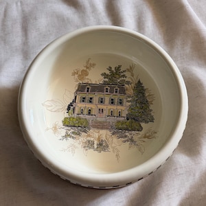 May include: A round, off-white ceramic bowl featuring a detailed illustration of a stately house with a green roof and surrounding trees. The bowl's rim has a decorative border, and the interior design includes floral accents.