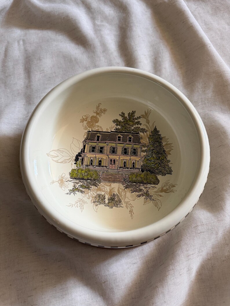 May include: A round, off-white ceramic bowl featuring a detailed illustration of a stately house with a green roof and surrounding trees. The bowl's rim has a decorative border, and the interior design includes floral accents.