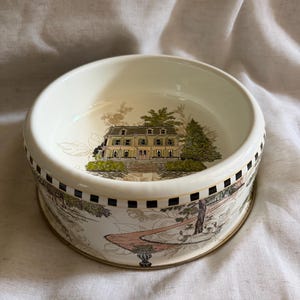 May include: Cream-colored ceramic bowl with a detailed landscape design. The bowl features a yellow building with green trees and a pathway. The rim has a black and white checkered pattern and a gold trim.
