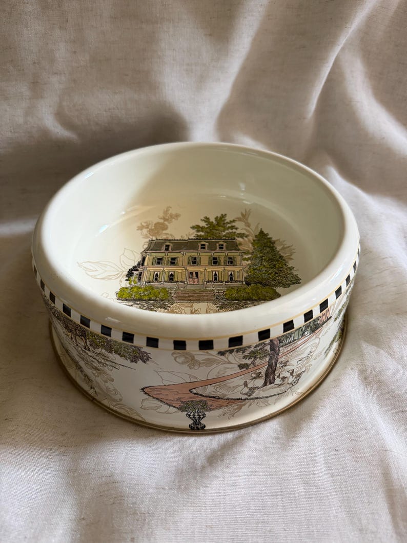 May include: Cream-colored ceramic bowl with a detailed landscape design. The bowl features a yellow building with green trees and a pathway. The rim has a black and white checkered pattern and a gold trim.
