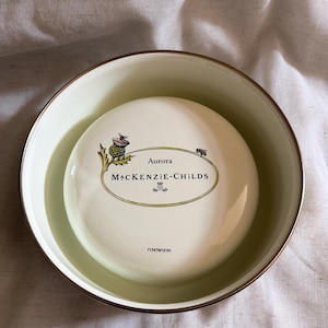 May include: A white ceramic bowl with a gold rim, containing a smaller white ceramic piece. The smaller piece has the text "Aurora MACKENZIE-CHILDS" in an oval design, with a thistle illustration. The bowl is filled with a light green liquid.
