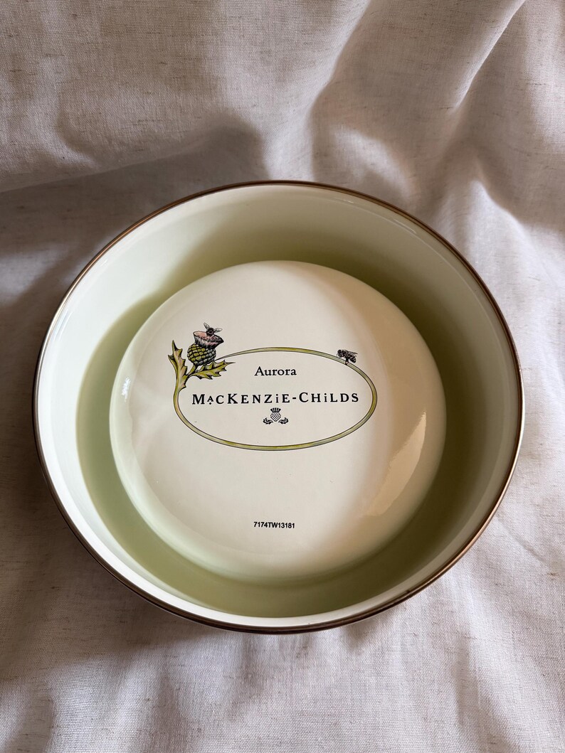 May include: A white ceramic bowl with a gold rim, containing a smaller white ceramic piece. The smaller piece has the text "Aurora MACKENZIE-CHILDS" in an oval design, with a thistle illustration. The bowl is filled with a light green liquid.