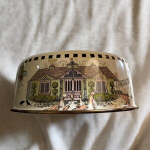 May include: A cream-colored, dome-shaped decorative bowl with a detailed illustration of a house with a pink roof and green walls. The bowl has a gold rim and a black and gold patterned border. The scene includes birds and other decorative elements.