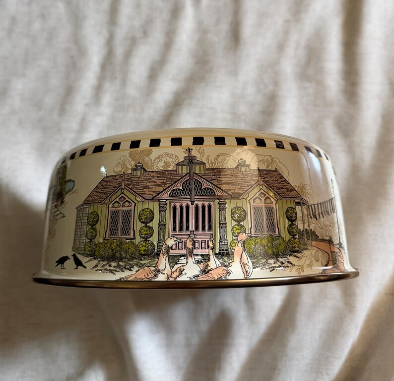 May include: A cream-colored, dome-shaped decorative bowl with a detailed illustration of a house with a pink roof and green walls. The bowl has a gold rim and a black and gold patterned border. The scene includes birds and other decorative elements.