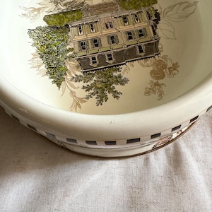 May include: A cream-colored, vintage-style bowl with a detailed illustration of a house surrounded by trees. The bowl's rim features a checkered pattern with gold accents, and floral designs. The overall aesthetic is reminiscent of antique tableware.